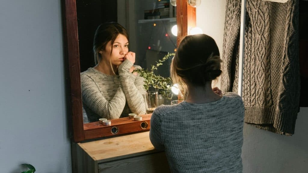 A woman in a grey sweater looks into a lighted mirror in a dim room.