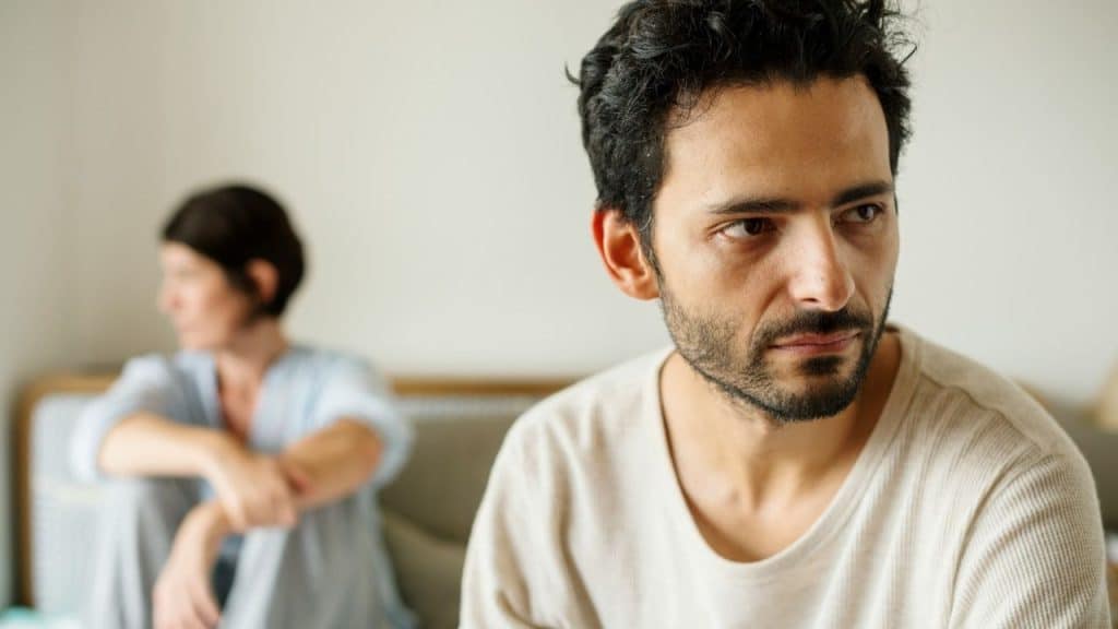 A bearded man looks away with a serious expression while a woman sits blurred behind him.