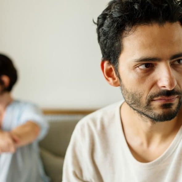 A bearded man looks away with a serious expression while a woman sits blurred behind him.