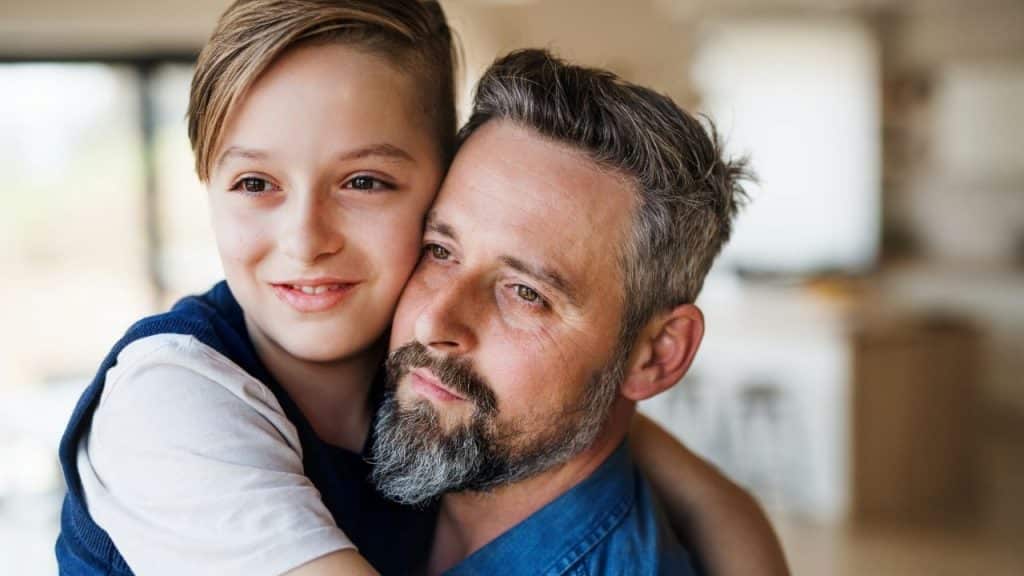 A young boy hugs a man with a graying beard from behind while both look away.