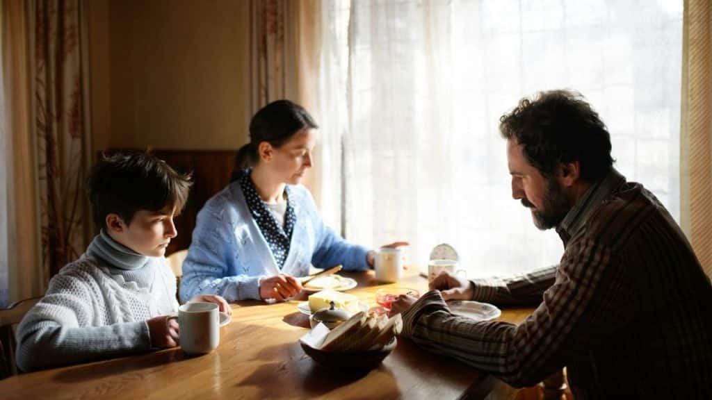 A man, woman, and child sit at a wooden table looking down during a meal.