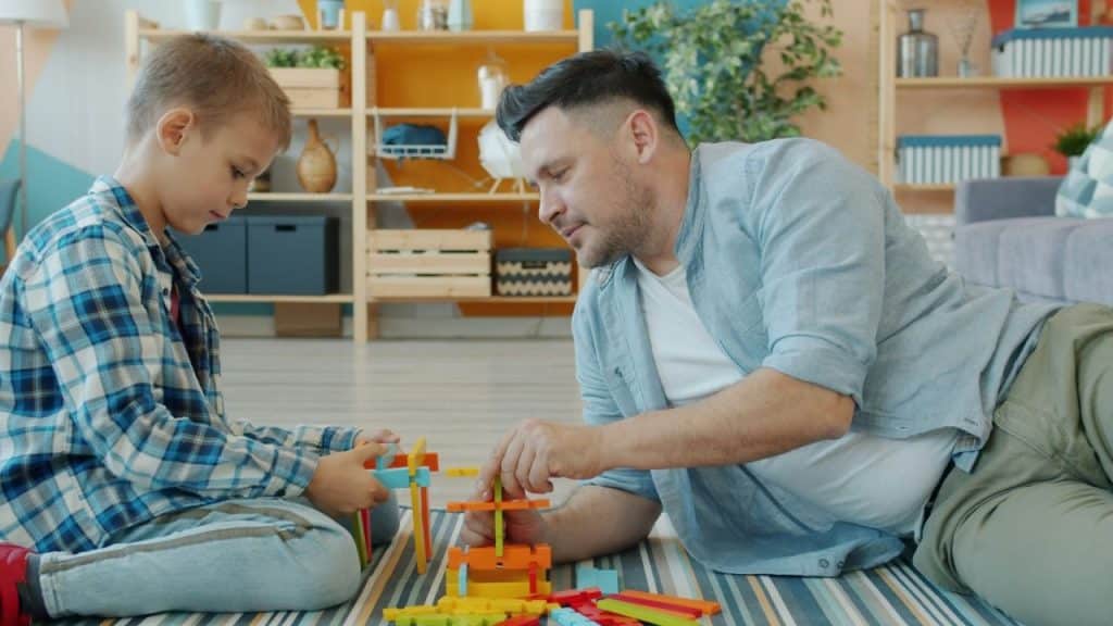 A man and a young boy lie on a striped rug and play with colorful blocks.