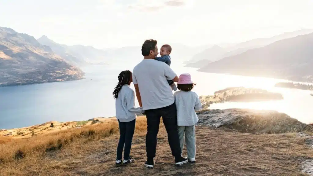 A man stands with three children overlooking a large lake and mountains during a sunset.