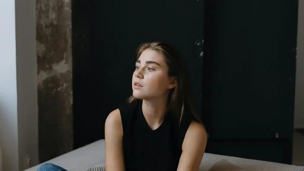 A thoughtful young woman sits on a bed looking off to the side.