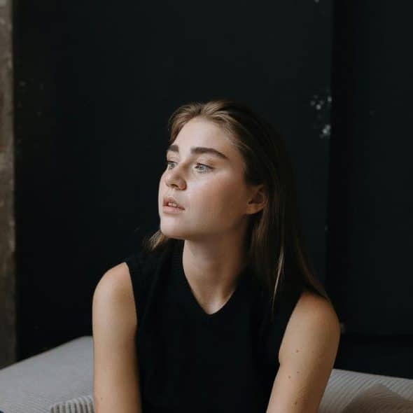 A thoughtful young woman sits on a bed looking off to the side.