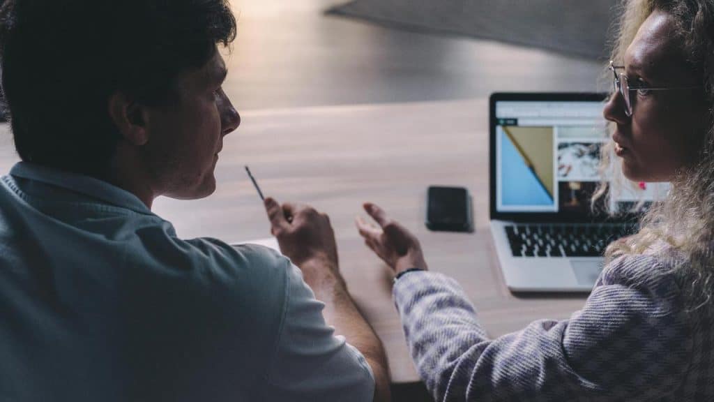 A man and a woman discussing work at a table with a laptop open.