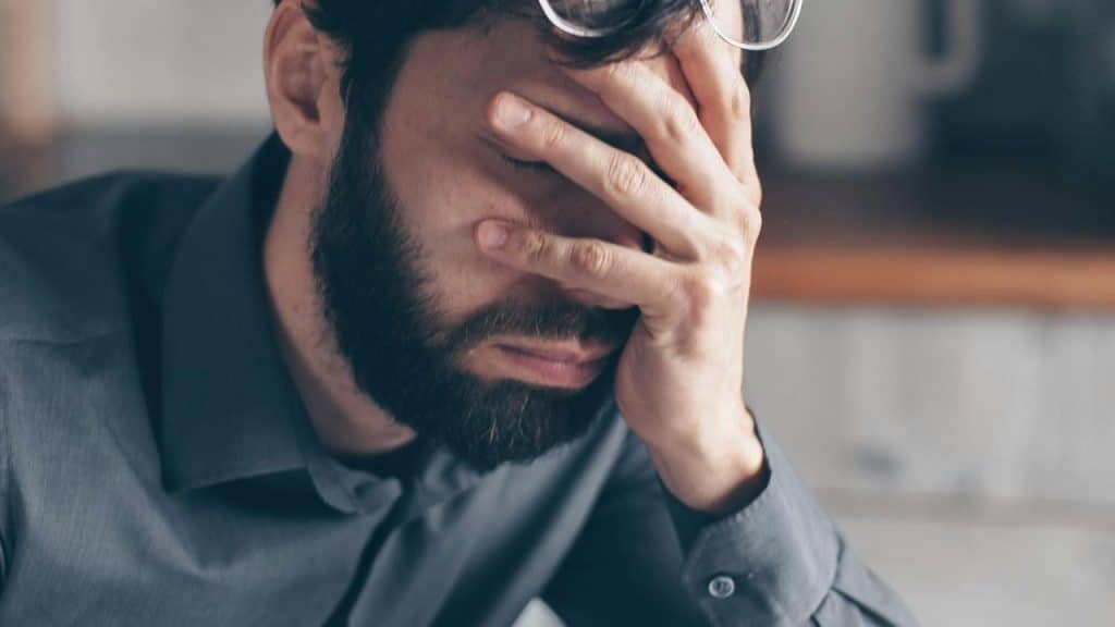 A stressed man with a beard covers his face with his hand while sitting indoors.