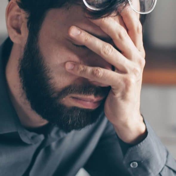 A stressed man with a beard covers his face with his hand while sitting indoors.