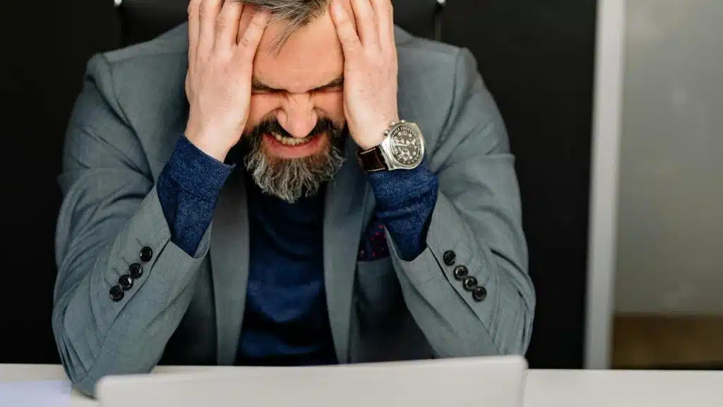 A frustrated businessman in a suit holds his head while sitting at a laptop.