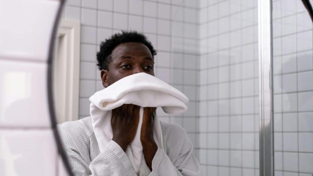 A person standing in a bathroom holding a white towel to their face.