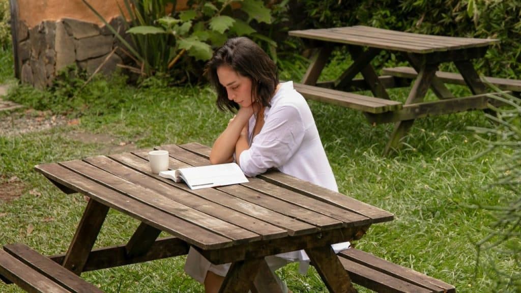 A woman sits alone at an outdoor wooden picnic table reading a book with coffee.