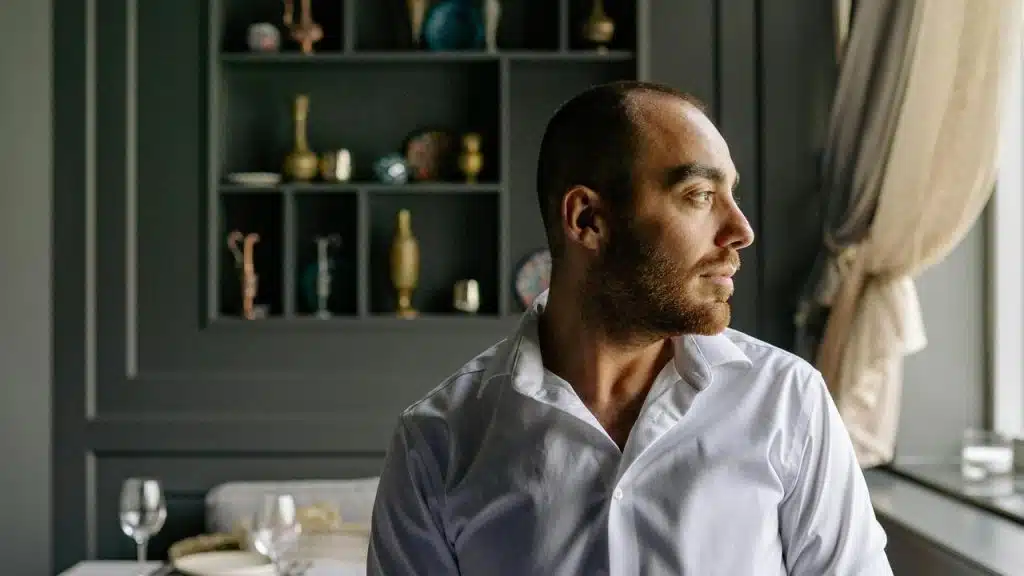 A man in a white shirt looking out of a window thoughtfully.