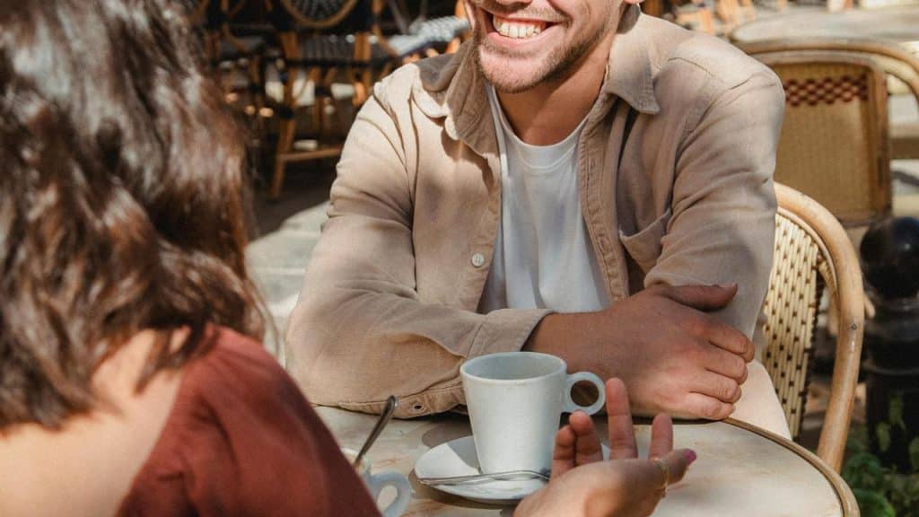 A man smiling while having coffee with someone at an outdoor café.
