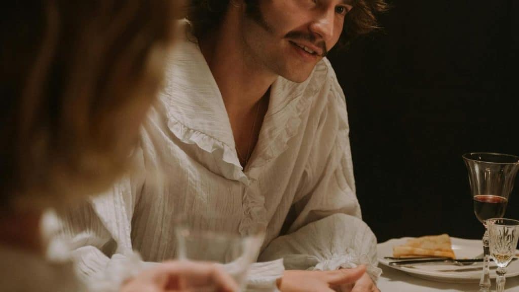 A man in a ruffled white shirt talking at a dinner table with wine glasses.