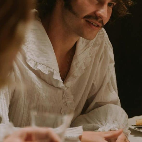 A man in a ruffled white shirt talking at a dinner table with wine glasses.
