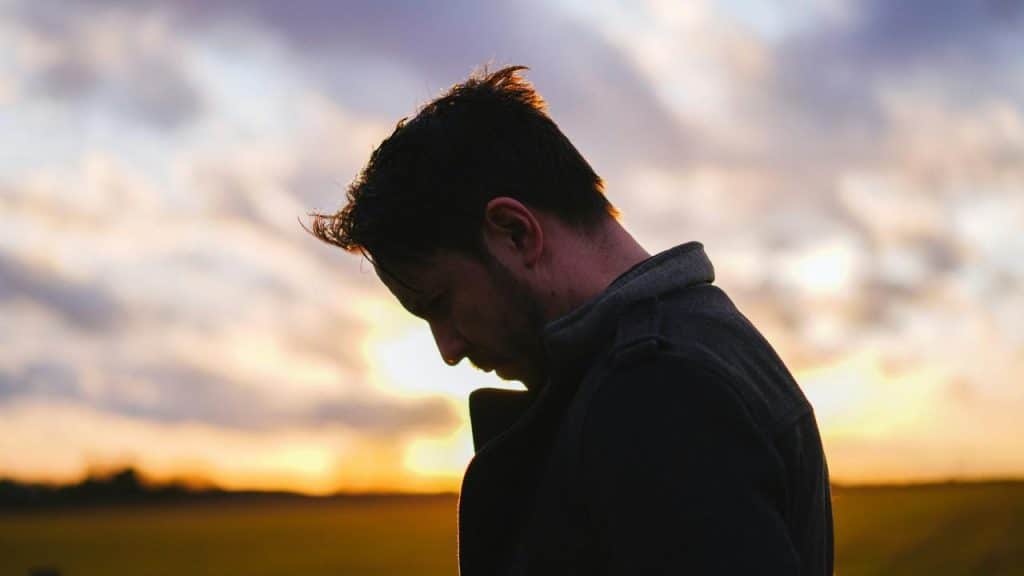 A man looks downward in silhouette against a colorful sunset sky.