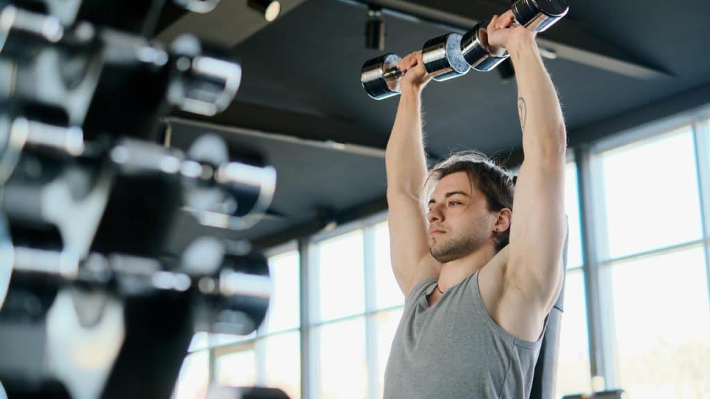 A man lifting dumbbells overhead while exercising in a gym.
