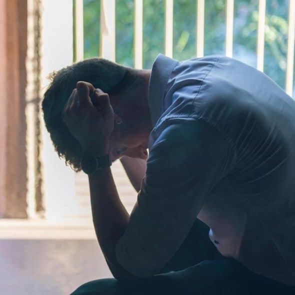 A man sitting with his head bowed and hands clasped in distress near a doorway.