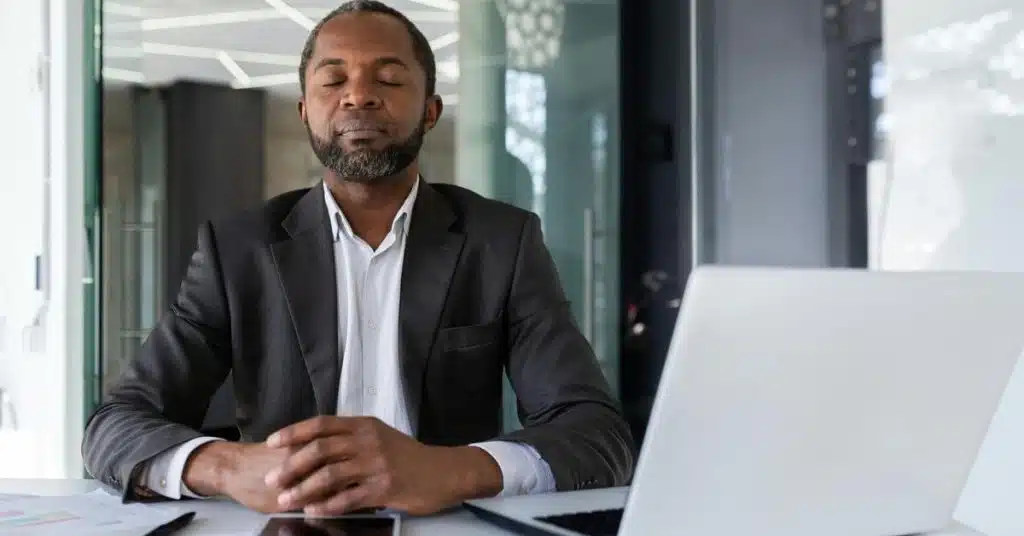 A man in a business suit sitting at a desk with a laptop, eyes closed as if meditating.