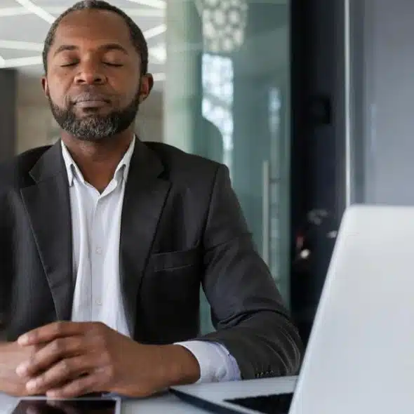 A man in a business suit sitting at a desk with a laptop, eyes closed as if meditating.