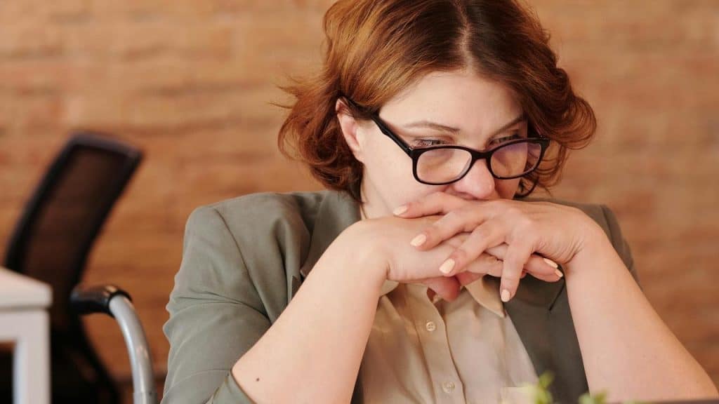 A worried woman wearing glasses with her hands clasped near her face.