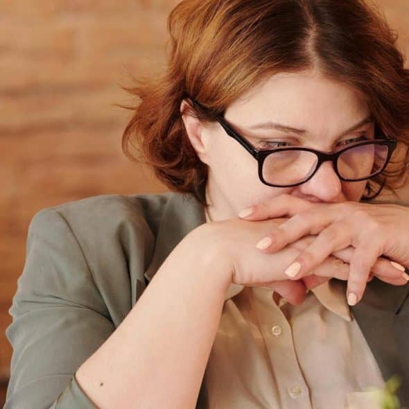 A worried woman wearing glasses with her hands clasped near her face.