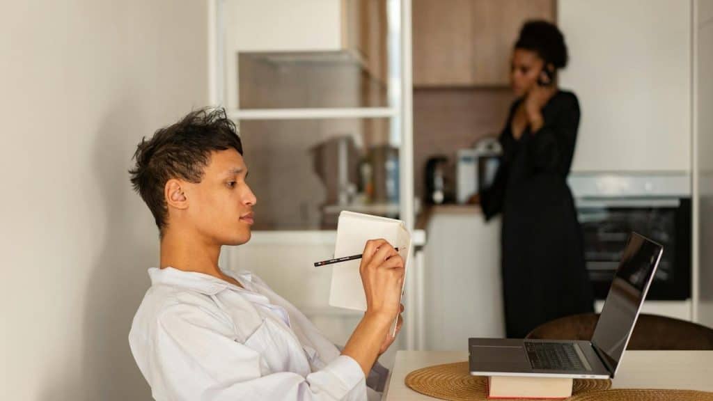 A man writing in a notebook at a table while a woman talks on the phone in the background.