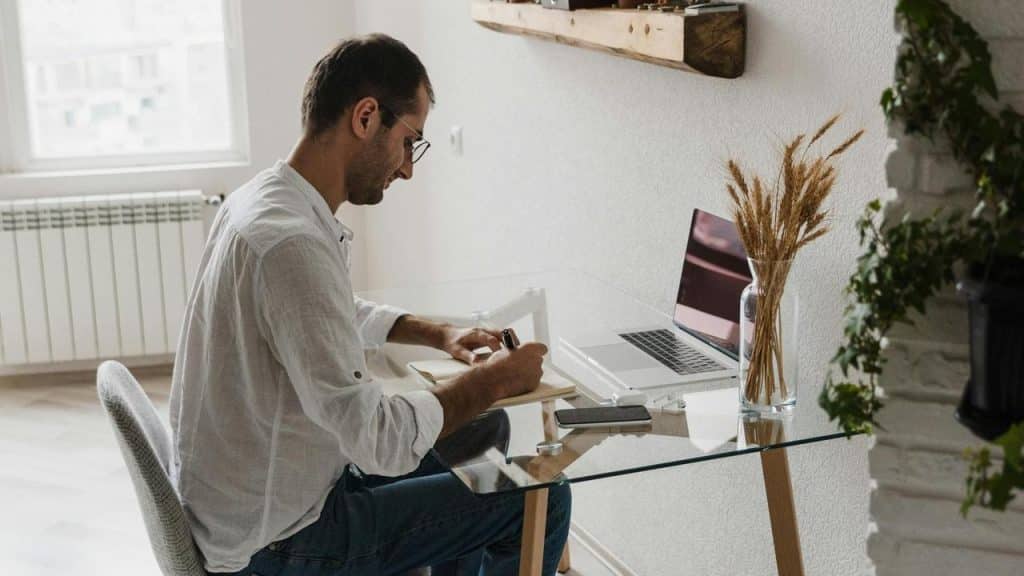 A man writing on a notebook while working on a laptop.