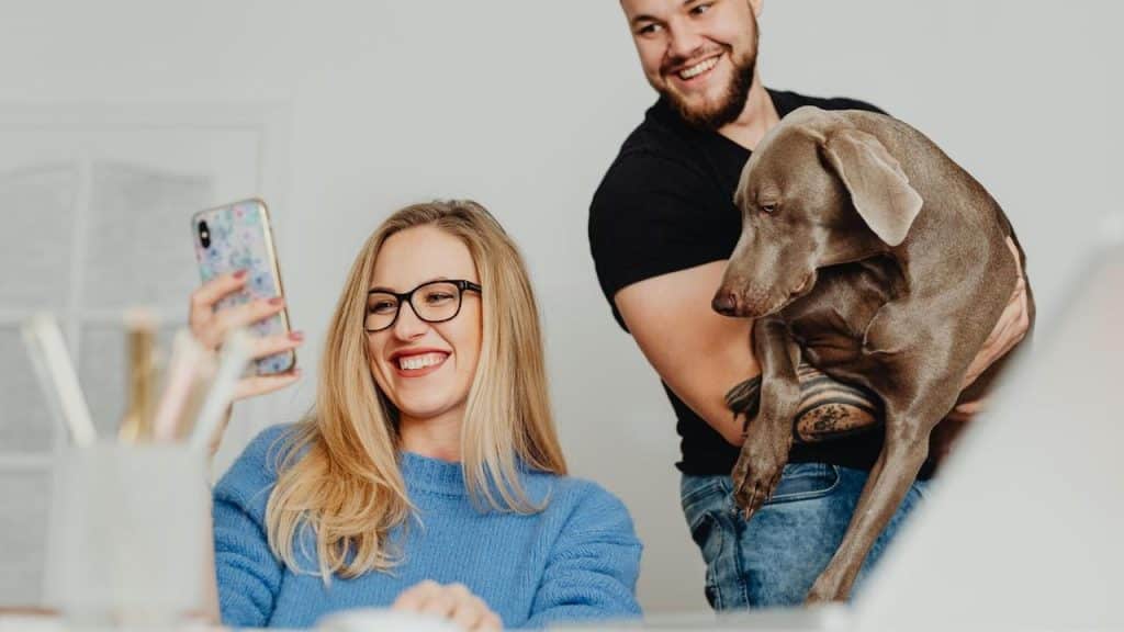 A woman smiling at her phone while a man holds their dog behind her.