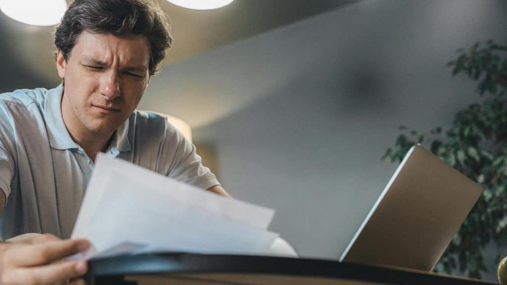 A man looking at documents with a concerned expression beside a laptop.