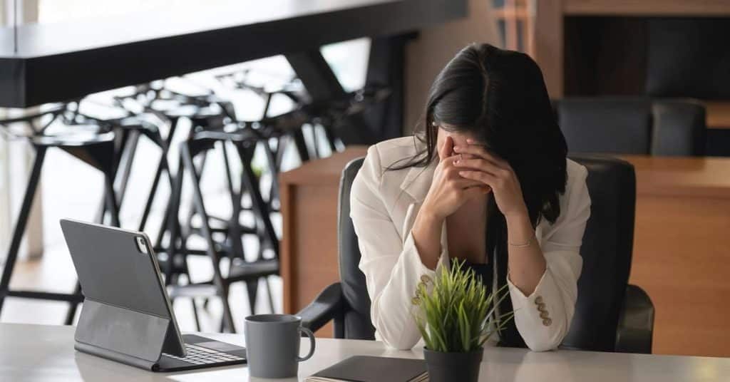 A woman sitting at a desk with her head in her hands beside a tablet and coffee cup.