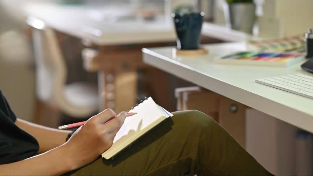 A person sits at a desk writing in a notebook with a pencil.
