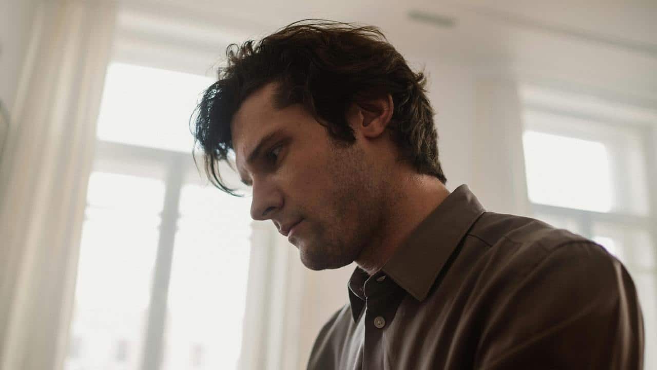 A man in a brown shirt looking down with a thoughtful expression near a bright window.
