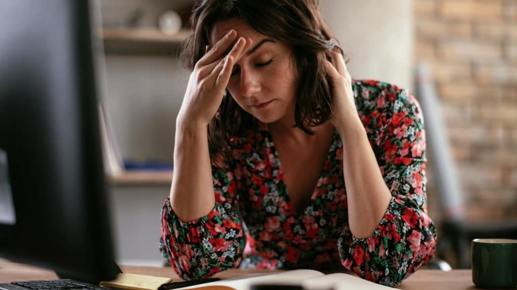 A woman sits at a desk with her hand on her forehead, looking down at work materials.