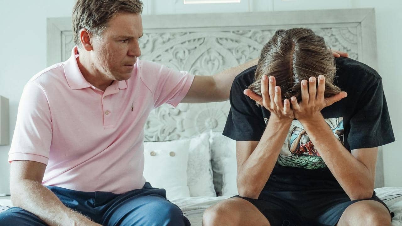 A father comforting his son, who is sitting with their face in their hands.