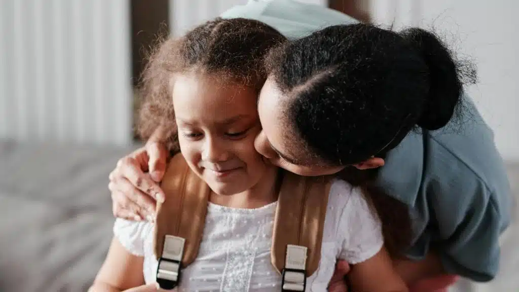 A mother hugging and kissing her smiling daughter wearing a backpack.