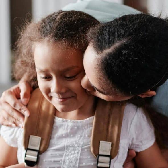 A mother hugging and kissing her smiling daughter wearing a backpack.