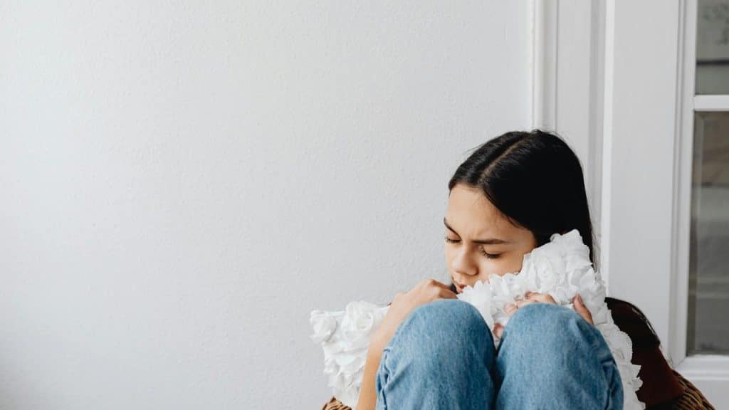 A sad young woman sitting curled up and hugging a pillow in a corner.