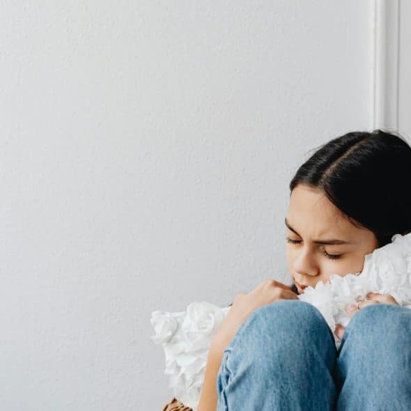 A sad young woman sitting curled up and hugging a pillow in a corner.