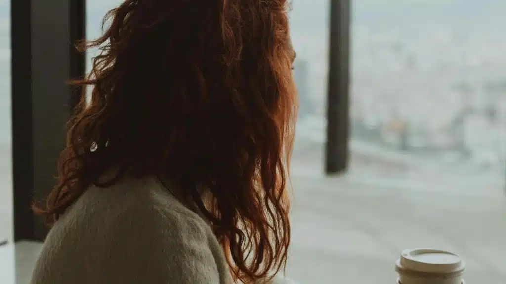 A person with wavy red hair looking out a window while holding a coffee cup.