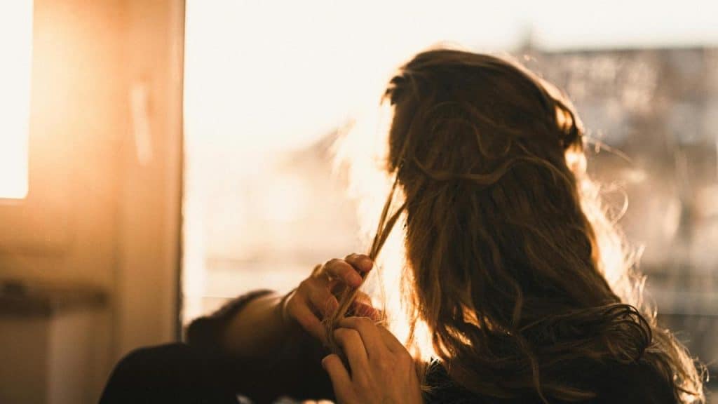 A person sitting by a sunlit window, twirling their hair in their fingers.