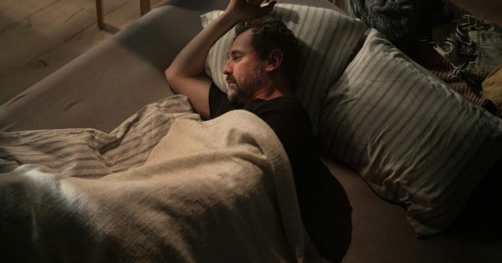 A man lying awake in bed under a blanket at night.