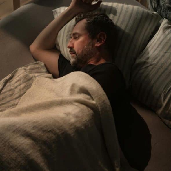 A man lying awake in bed under a blanket at night.