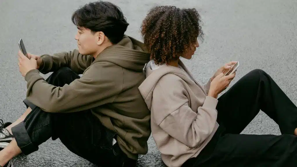 A pair of people sitting back-to-back on the ground, each focused on their phone.