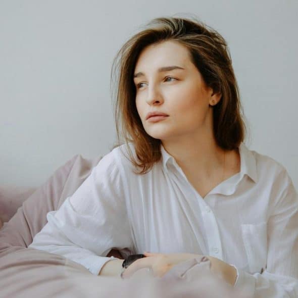 A thoughtful woman in a white shirt sitting on a bed and looking away.
