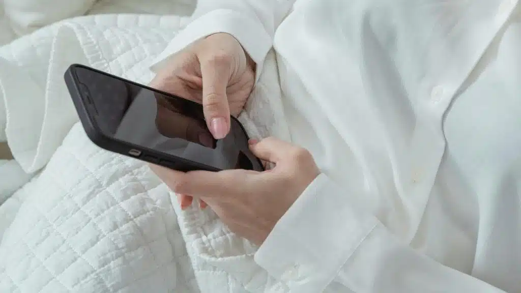 A person in a white shirt holding a smartphone while sitting on a bed.