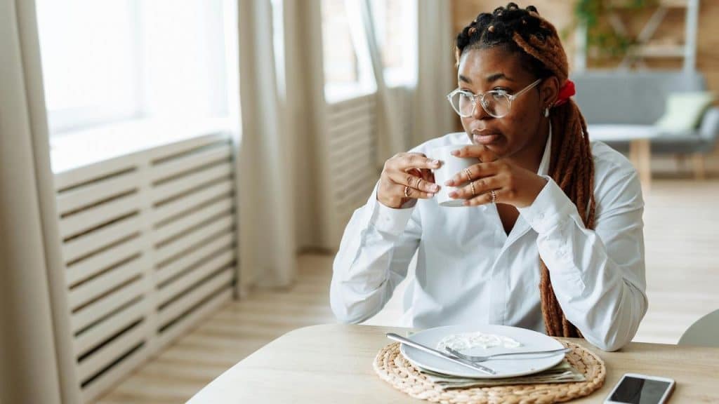 A woman wearing glasses sitting at a table and holding a cup in a bright room.
