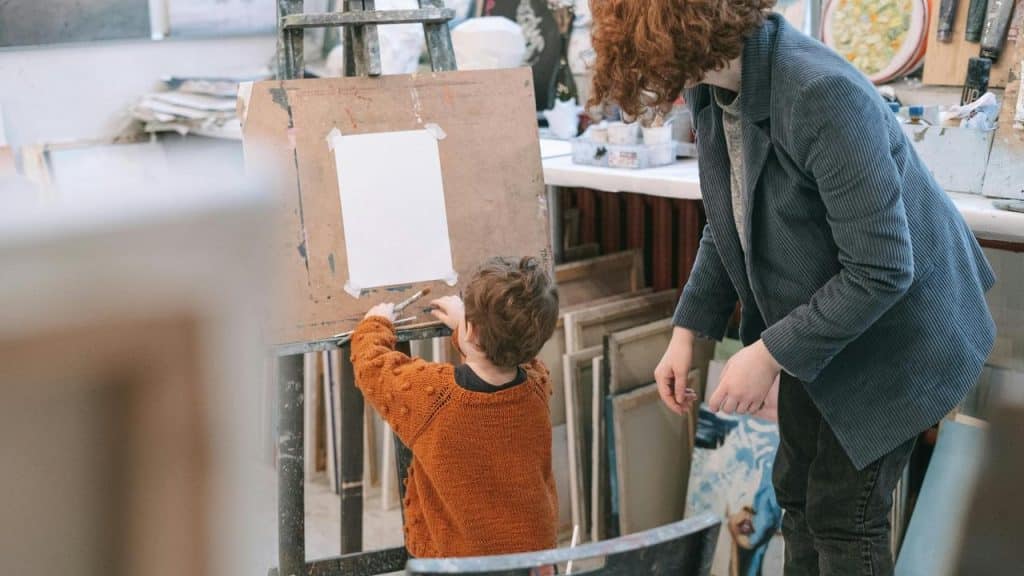 A child drawing on a paper at an easel with an adult assisting nearby.