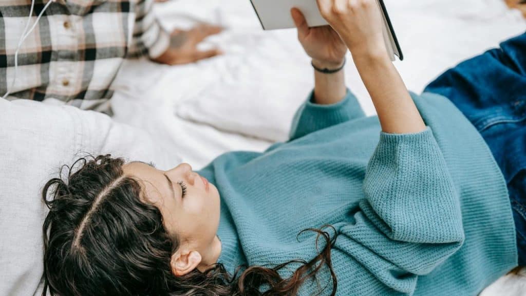 A woman lying on a bed reading a book while wearing a blue sweater.