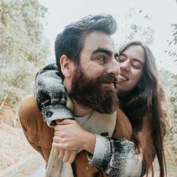 A man with a beard and a brown jacket gives a piggyback ride to a smiling woman with long brown hair along a wooded path.
